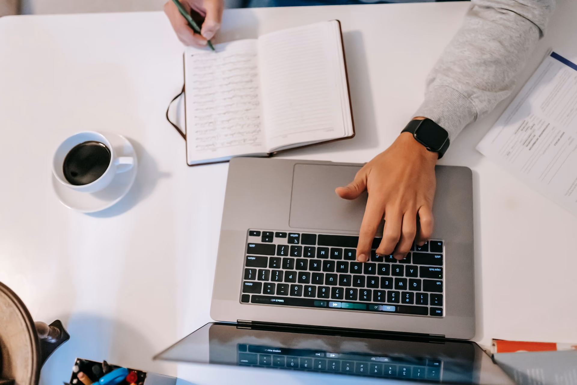 Overhead view of hands writing in a notebook beside an open laptop and coffee