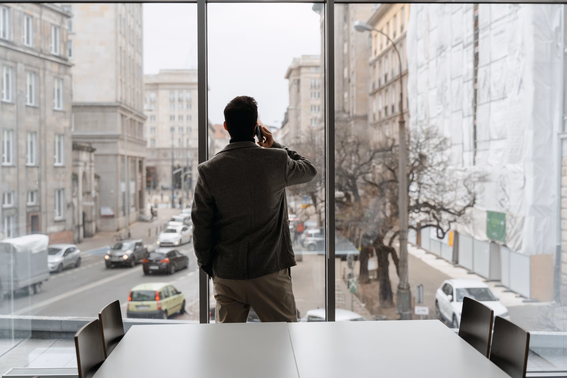 A man in a blazer on a phone call, looking out over a city street from an office window